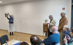 Eine der Künstlerinnen steht vorne im Saal und hält einen Eimer hoch. Sie trägt eine Maske.&nbsp;Rechts steht ein weiterer Künstler, der Drehorgel spielt.&nbsp;