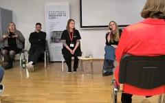 Katrin beantwortet eine Frage aus dem Publikum mit Mikrofon in der Hand. Neben ihr sitzt Miriam. Auf der linken Seite sitzen die beiden Gebärdensprachedolmetscher.