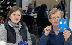 Manuela und Klaus beim Basteln. Klaus hält seine gebastelte Karte hoch. 