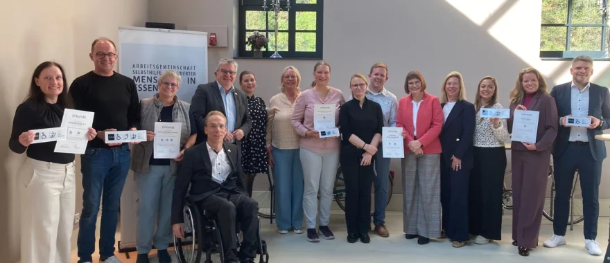 Die Betreiber*innen der ausgezeichneten Gebäude sowie Angela Ströter (AGSBM), Ralf Bockstedte (Inklusionsbeirat), Peter Renzel und Eva Koch (Stadt Essen) lächeln in Richtung Kamera. Die Betreiber*innenhalten ihre Signet-Urkunden und Schilder in der Hand.&nbsp;